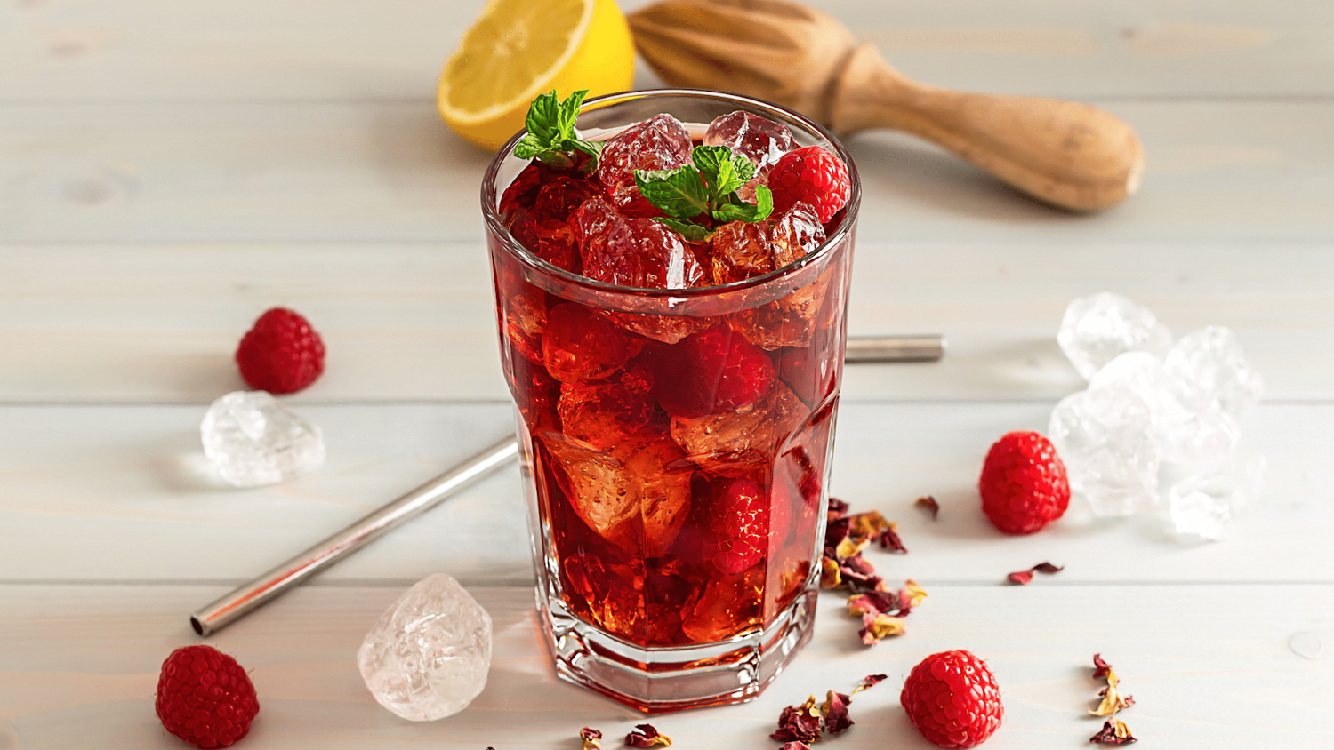 A Glass Of Homemade Iced Tea On A Table With Raspberries, Lemon, Ice, Herbs, And A Muddler